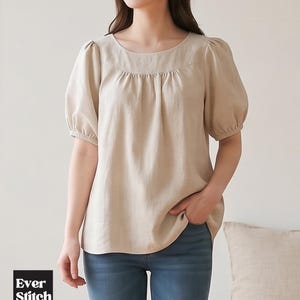 Può includere: Una camicetta beige a maniche corte con scollo rotondo, dettagli arricciati sul petto e maniche a sbuffo con polsini elasticizzati. Il capo è abbinato a un paio di jeans blu. È visibile un logo quadrato nero con il testo Ever Stitch.