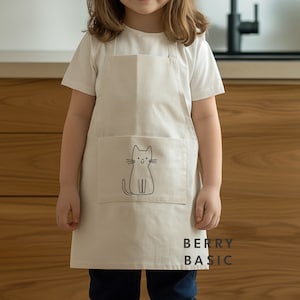 Op de afbeelding: Een crèmekleurig schort met een zwarte kattenillustratie op de voorzak. Het schort wordt door een persoon gedragen en heeft een eenvoudig ontwerp. De tekst "BERRY BASIC" staat in het zwart rechtsonder op het schort.