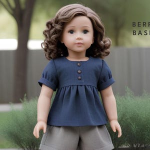 Può includere: Una bambola che indossa una camicetta blu navy a maniche corte con vita arricciata e tre bottoni marroni sul davanti. La bambola indossa anche pantaloncini grigi. Il testo "BERRY BASIC" è visibile nell'angolo in alto a destra dell'immagine.