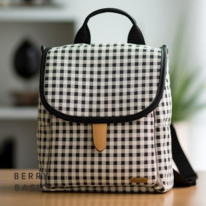 May include: A black and white gingham patterned diaper bag with a black leather trim and a brown leather handle. The bag has a zipper closure and a small pocket on the front.