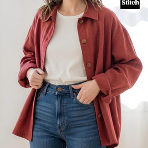 May include: A rust-colored button-down shirt with large wooden buttons, worn over a white t-shirt and paired with blue jeans. The shirt has long sleeves and a relaxed fit. The jeans are a classic blue wash.