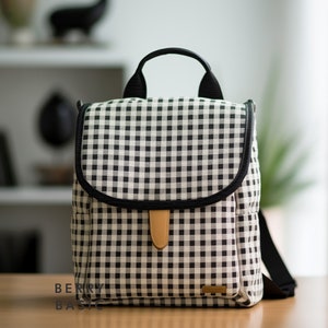 May include: A black and white checkered backpack with a brown leather handle and a black strap. The backpack has a black trim around the edges and a small brown leather patch on the front.