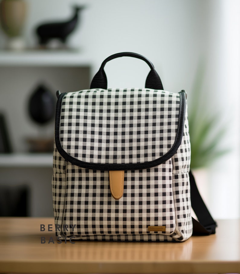 May include: A black and white checkered backpack with a brown leather handle and a black strap. The backpack has a black trim around the edges and a small brown leather patch on the front.