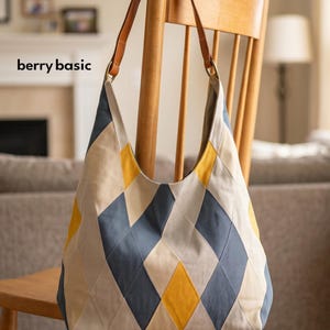 Peut inclure: Un sac hobo arborant un motif géométrique argyle dans des tons bleu ardoise, jaune moutarde et beige. Le sac possède une bandoulière en cuir marron fixée par des anneaux métalliques et le texte berry basic imprimé en noir sur le côté gauche.