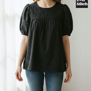 Puede incluir: Top negro de manga corta con cuello redondo y detalles bordados. El top tiene un corte holgado y mangas abullonadas. La marca "Ever Stitch" está en un recuadro negro. La modelo lleva vaqueros azules.