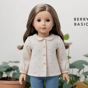 May include: A doll with long brown hair and blue eyes wearing a cream blouse with small pink floral patterns and blue jeans. The blouse has a rounded collar and button-up front. The text "BERRY BASIC" is visible.