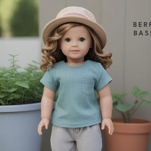 May include: A doll wearing a teal blue t-shirt with a pocket and a brown hat. The doll has long brown hair and blue eyes. The doll is wearing beige trousers. The text "BERRY BASIC" is visible in the background.