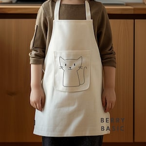 Puede incluir: Delantal color crema con una ilustración de gato negro en el bolsillo delantero. El delantal tiene correas blancas y el texto "BERRY BASIC" impreso en la esquina inferior derecha. El delantal lo lleva una persona.