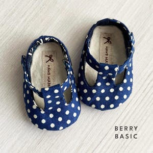 Könnte beinhalten: Ein Paar blaue Babyschuhe mit weißen Punkten. Die Schuhe haben einen T-Riemen und sind handgefertigt. Das Etikett auf den Schuhen lautet "HAND MADE".