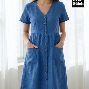 Op de afbeelding: Blauwe linnen jurk met een V-hals, korte mouwen en een knoopsluiting aan de voorkant. De jurk heeft twee voorzakken en een getailleerde taille. Het Ever Stitch-logo is rechtsboven zichtbaar.