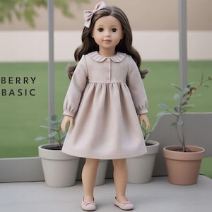Könnte beinhalten: Eine Puppe trägt ein hellbraunes Kleid mit einem weißen Kragen und rosa Knöpfen. Das Kleid hat lange Ärmel und einen gerafften Rock. Die Puppe hat braunes Haar und blaue Augen und trägt eine rosa Schleife im Haar. Die Puppe steht auf einem Holzboden mit grünem Hintergrund.