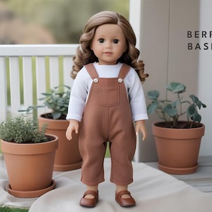 May include: A doll wearing a white long-sleeved shirt and brown overalls with a single front pocket. The doll has long brown hair and brown eyes. The doll is standing on a wooden surface in front of a white picket fence and potted plants.