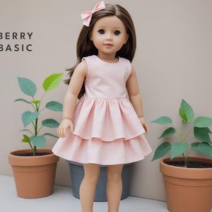 May include: A pink dress with a ruffled skirt and a bow in the hair of a doll. The doll is standing in front of a light brown wall with two potted plants.