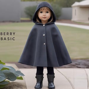 May include: A dark gray hooded cape with button closures. The cape is worn by a child with long blonde hair. The child is standing on a stone path with green grass in the background.