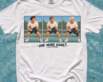 以下が含まれることがあります： 白い半袖Tシャツ。コートでピックルボールをしながら年齢を重ねていく人物を描いた3つのパネルのグラフィックプリントが施されている。パネルの下には、黒い大文字で「ONE MORE GAME?」というテキストと、その下に水平線が描かれている。
