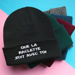 Peut inclure: Un ensemble de bonnets en tricot noir, vert, bordeaux et gris. Le bonnet du dessus est brodé en blanc avec l'inscription "QUE LA RACLETTE SOIT AVEC TOI".