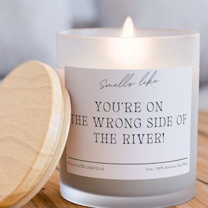 以下が含まれることがあります： 木製のテーブルの上に、木製の蓋が付いた、火の灯ったすりガラスのキャンドルがあります。キャンドルには「Smells like You're on the Wrong Side of the River！」というテキストが書かれた白いラベルが付いています。