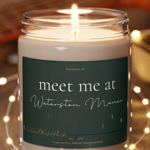 May include: A brown candle with a green label that reads "Essence of meet me at Waterston Manor". The candle is sitting on a wooden coaster and is surrounded by string lights.