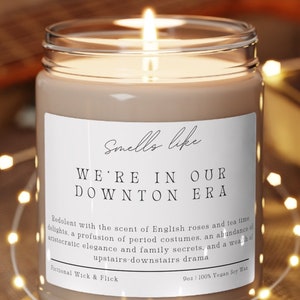 May include: A lit candle in a glass jar with a white label that reads "Smells like WE'RE IN OUR DOWNTON ERA". The candle sits on a wooden coaster, surrounded by string lights. The background includes a guitar and a book.