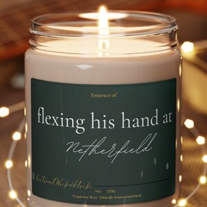 Könnte beinhalten: Eine Kerze mit einem grünen Etikett, auf dem "Essence of flexing his hand at Netherfield" steht. Die Kerze ist angezündet und steht auf einem Holzuntersetzer.