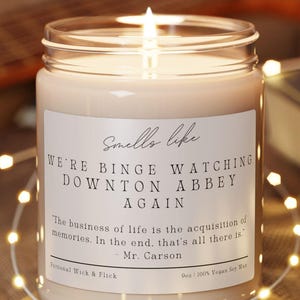 May include: A lit candle in a glass jar with a label that reads "Smells like We're Binge Watching Downton Abbey Again." The candle sits on a round wooden coaster. A string of fairy lights and a guitar are in the background.