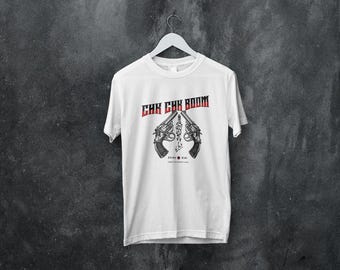 May include: White t-shirt with the text "CHK CHK BOOM" in red and a graphic of two revolvers with a chain and the text "Stray Kids" below. The shirt is hanging on a white hanger.