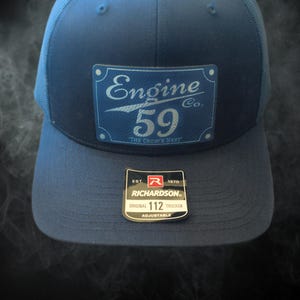 Può includere: Un cappello da camionista blu con una rete bianca sul retro. Il cappello presenta una toppa blu e bianca con il testo "Engine 59 Co. The Crow's Nest". Il cappello ha anche un'etichetta nera e rossa con il testo "Richardson Original 112 Trucker Adjustable".