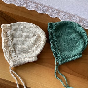 May include: Two knitted baby bonnets, one in cream and one in green, with ties. The bonnets have a lacy design around the edge.