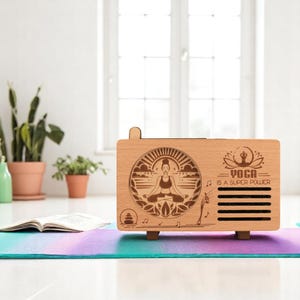K&ouml;nnte beinhalten: Holzkiste mit einem Laser-geschnittenen Design einer Person, die im Lotussitz meditiert. Die Kiste hat eine helle Holzoptik und der Text "Yoga is a super power" ist auf der Vorderseite eingraviert.