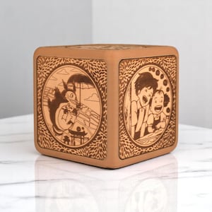 May include: A square, light brown wooden box with engraved illustrations from a Japanese animated film. The box features detailed circular designs with characters and scenes, surrounded by a leaf pattern.