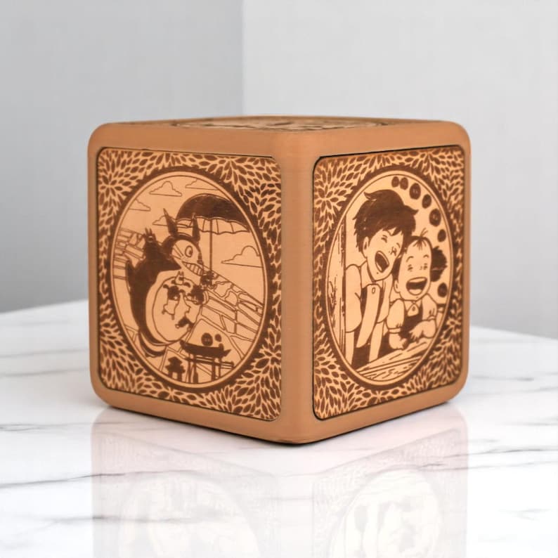 May include: A square, light brown wooden box with engraved illustrations from a Japanese animated film. The box features detailed circular designs with characters and scenes, surrounded by a leaf pattern.
