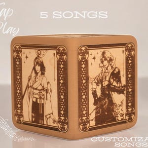 Peut inclure: Boîte à musique cubique en bois avec des illustrations gravées de style anime. Le texte "Tap to Play", "5 SONGS" et "CUSTOMIZABLE SONGS" est visible. La boîte a une finition marron clair et une bordure décorative.