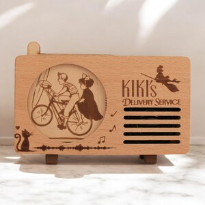 May include: A wooden music box with a laser-engraved image of Kiki and her friend riding a bicycle. The music box has a speaker grill and the text "Kiki's Delivery Service" on the front.