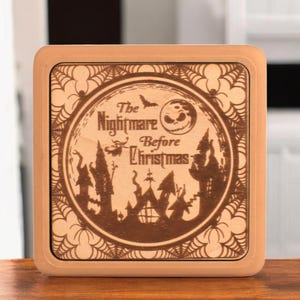 May include: A wooden plaque with a brown background and a white border. The plaque features a circular design with a black and white image of Jack Skellington from the movie "The Nightmare Before Christmas". The text "The Nightmare Before Christmas" is written in a black font.