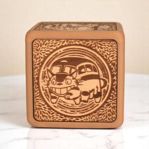 May include: A square wooden box with a detailed laser-etched design. The central image features a smiling cartoon character in a bus, surrounded by a circular frame. The box has a light brown color with a decorative leaf pattern.