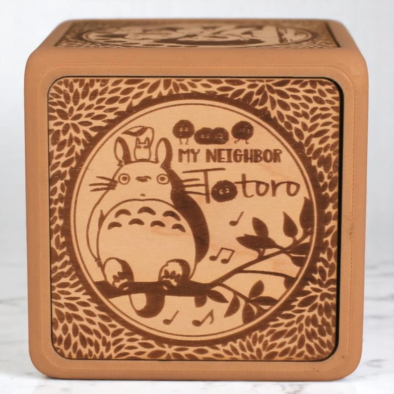 May include: A square wooden music box with a light brown frame and a laser-etched design. The design features the words "My Neighbor Totoro" and an illustration of Totoro, a character from the animated film, surrounded by leaves and musical notes.