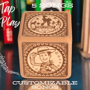 Peut inclure: Un coffret à musique en bois avec un motif brun et blanc représentant Woody de Toy Story. Le coffret à musique a un bouton qui dit "Tap to Play" et le texte "5 Songs" et "Customizable Songs".