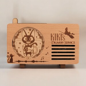 May include: A wooden music box shaped like a vintage radio with a laser-engraved design of Kiki's Delivery Service. The front panel features a black cat in a mug with stars and flowers. The text "Kiki's Delivery Service" is engraved on the right side of the box.