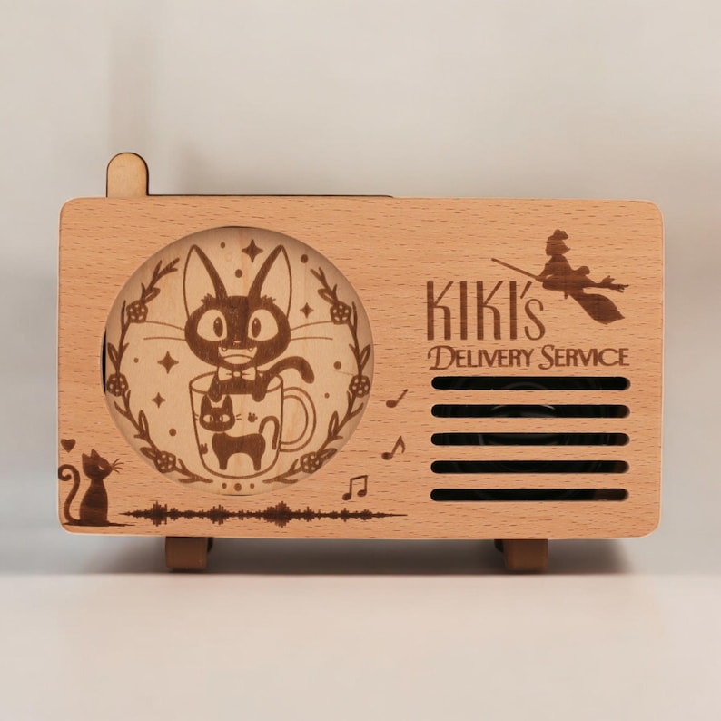 May include: A wooden music box shaped like a vintage radio with a laser-engraved design of Kiki's Delivery Service. The front panel features a black cat in a mug with stars and flowers. The text "Kiki's Delivery Service" is engraved on the right side of the box.