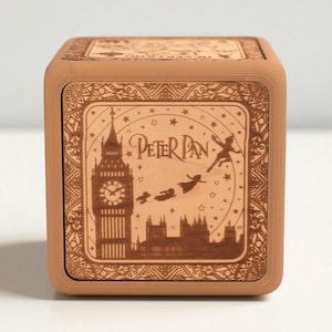 May include: A brown wooden music box featuring a laser-etched design of Peter Pan, Big Ben, and flying figures. The box has a square shape with a detailed border and the words "Peter Pan" in a decorative font.
