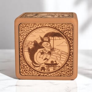 May include: A wooden music box with a light brown finish. The front panel features a circular engraved design of a cartoon character under an umbrella. The design is surrounded by a floral border. The top of the box also has an engraved design.