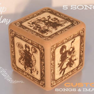 Könnte beinhalten: Ein Holzwürfel mit fünf Seiten, die mit gravierten Bildern von Menschen verziert sind. Die Oberseite des Würfels zeigt "5 SONGS" und die Vorderseite "Tap to Play". Die Unterseite des Würfels zeigt "CUSTOM SONGS & IMAGES".