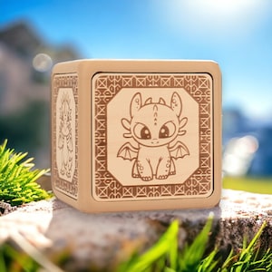May include: A wooden cube with a light brown finish features a decorative design of a cartoon dragon with large eyes. The dragon is outlined in a darker brown color and is surrounded by a geometric pattern.
