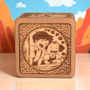 May include: A light brown wooden cube with a laser-etched design. The design features two laughing children inside a circular frame with a leafy border. The cube is set against a backdrop of a blue sky and orange mountains.