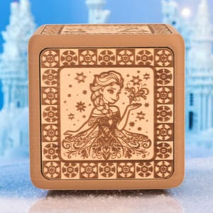 Könnte beinhalten: Eine Holz-Spieluhr mit kupferfarbener Oberfläche zeigt ein filigranes Design einer Prinzessin mit langen blonden Haaren und einem wallenden Kleid. Das Design ist von einem Rand aus Schneeflocken und anderen winterlichen Motiven umgeben.