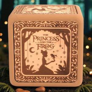 May include: A wooden box featuring a laser-cut design from "The Princess and the Frog." The box has a light brown finish with a detailed illustration of the princess, a frog, stars, and the moon. The title of the movie is also displayed.
