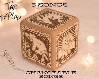 May include: A wooden cube music box with a Lion King theme. The cube features images of Simba and Mufasa. The text "Tap to Play" and "5 Songs" is printed on the box. The text "Changeable Songs" is printed below the box.