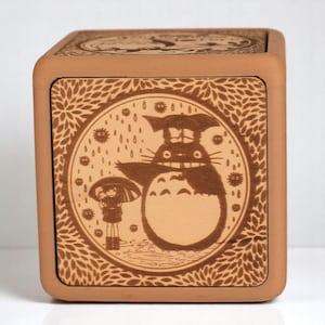 May include: A square, wooden box with a detailed, engraved design. The front panel features a circular illustration of Totoro from My Neighbor Totoro, with rain and a child under an umbrella. The box is a warm, earthy brown color.