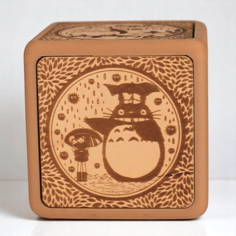 May include: A square, wooden box with a detailed, engraved design. The front panel features a circular illustration of Totoro from My Neighbor Totoro, with rain and a child under an umbrella. The box is a warm, earthy brown color.