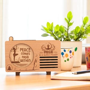 K&ouml;nnte beinhalten: Ein Holzlautsprecher mit einer hellen Holzoptik. Der Lautsprecher hat einen Ausschnitt mit einer Frau, die Yoga macht, mit dem Text "Peace comes from within" und "Yoga is a super power".